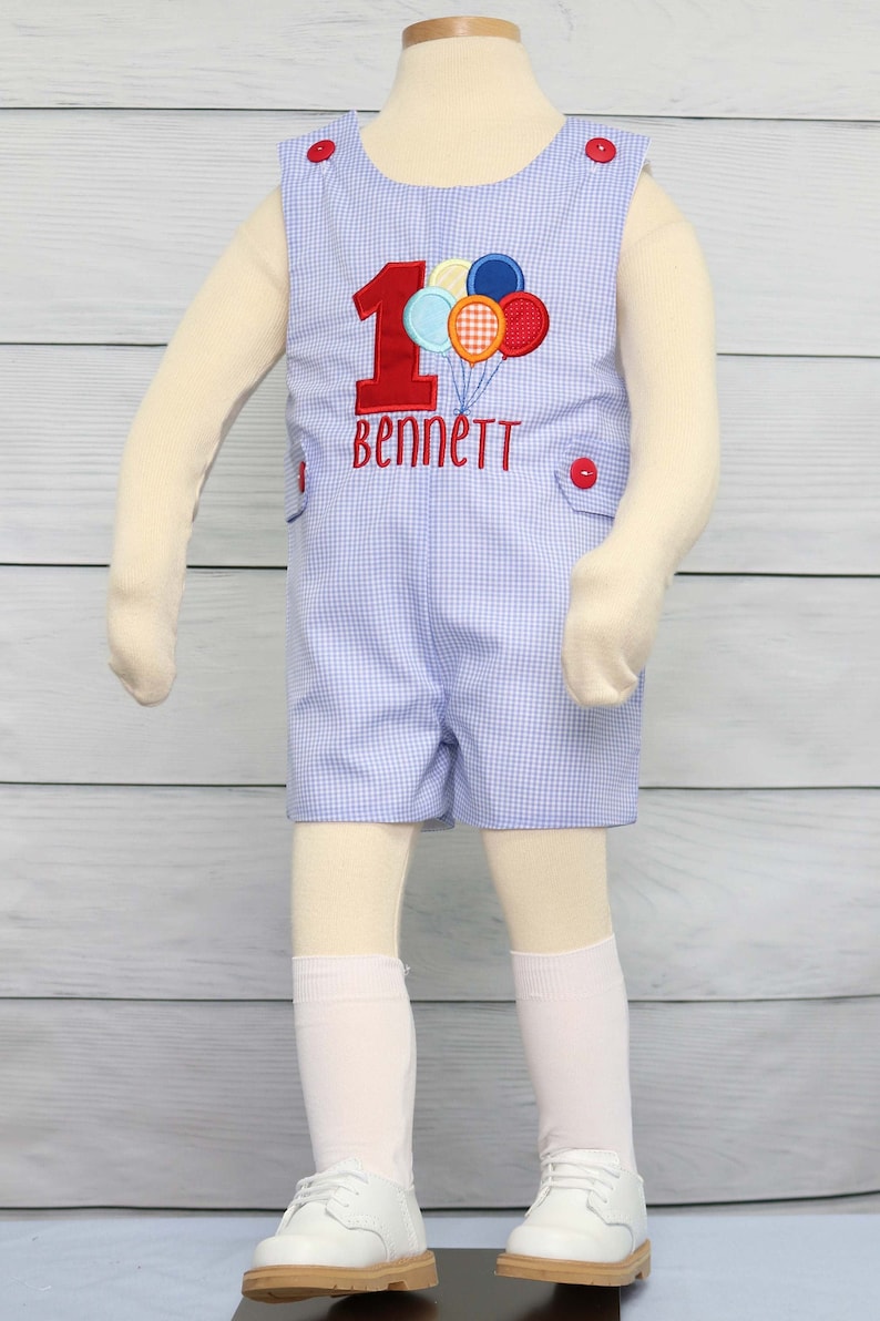 May include: A blue and white gingham romper with a red number one and colorful balloons embroidered on the front. The romper also has the name "Bennett" embroidered on the front.
