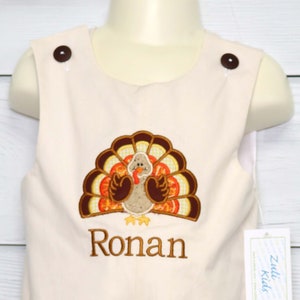 May include: A light beige sleeveless romper featuring two dark brown buttons at the shoulders. The front displays an embroidered turkey with brown, orange, and yellow feathers, and the name Ronan is embroidered in brown thread below the turkey.