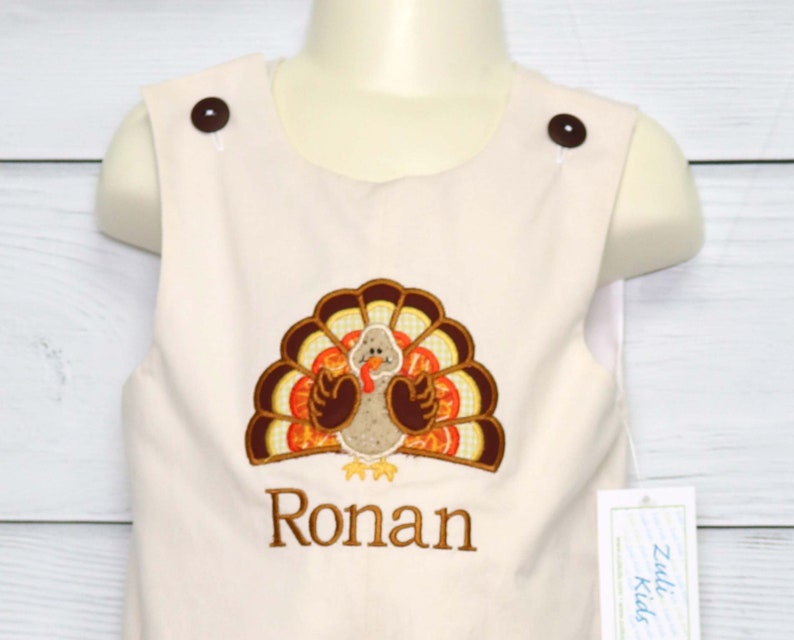 May include: A light beige sleeveless romper featuring two dark brown buttons at the shoulders. The front displays an embroidered turkey with brown, orange, and yellow feathers, and the name Ronan is embroidered in brown thread below the turkey.