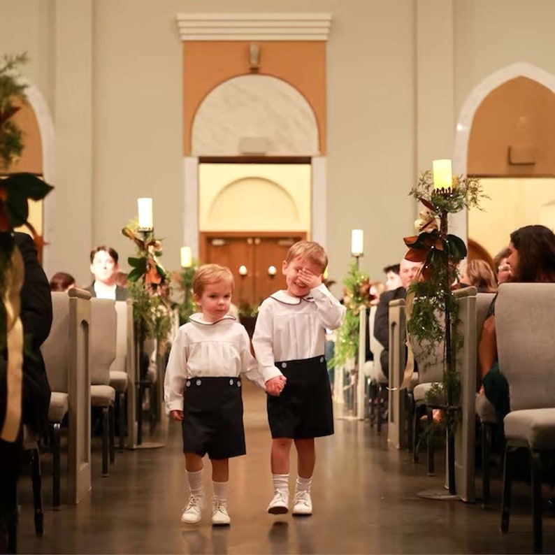 May include: Two young boys in matching outfits walk down an aisle. They wear white long-sleeved shirts with navy blue shorts, white socks, and white shoes. One boy covers his eyes with his hand. The setting appears to be a wedding ceremony.