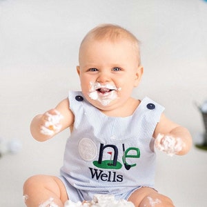 Peut inclure: Un bébé garçon portant une salopette rayée bleu et blanc avec un motif de golf "un" et le nom "Wells" brodé dessus. Le bébé est couvert de glaçage blanc et a une expression joyeuse sur son visage.