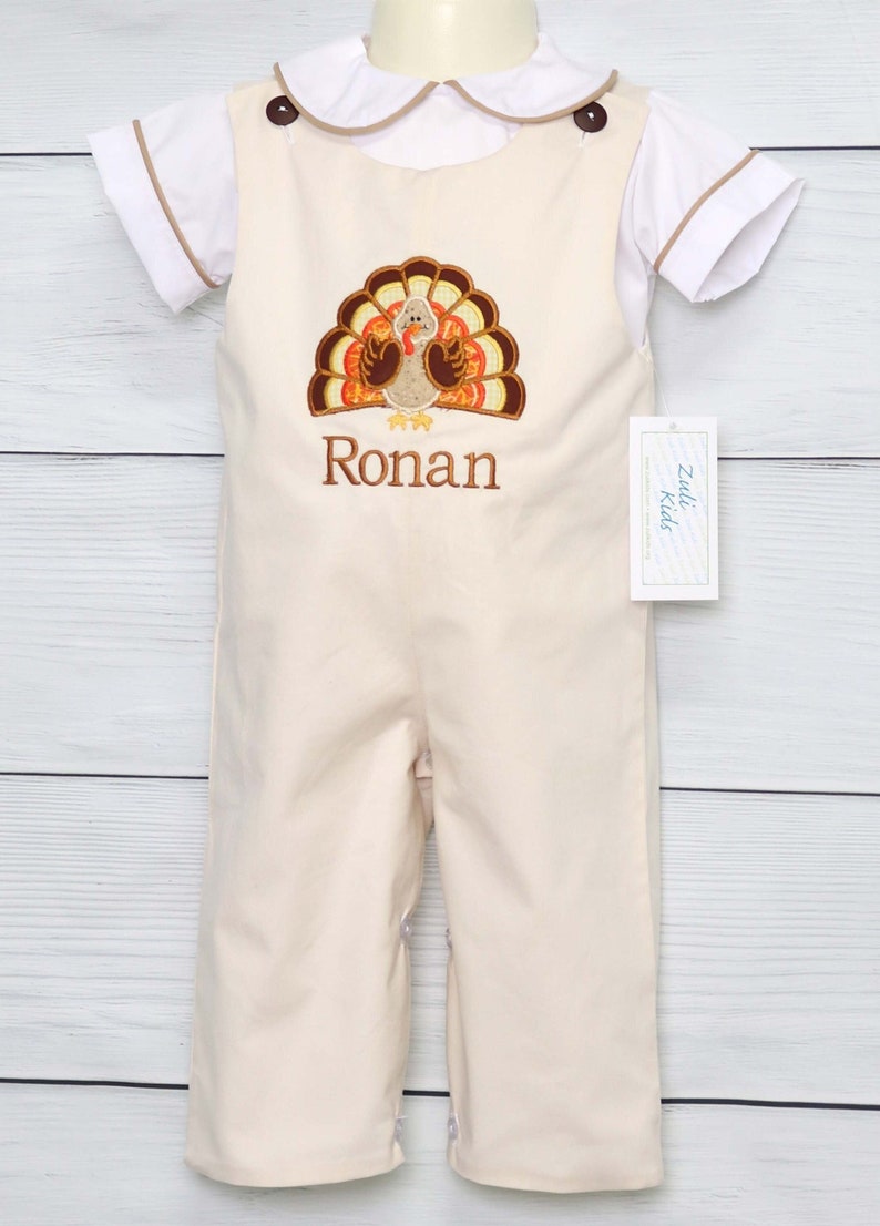 May include: A cream-colored overall with a white short-sleeved shirt featuring a Peter Pan collar. The overall has an embroidered turkey design and the name Ronan stitched in brown thread. Two dark brown buttons secure the straps at the shoulders.