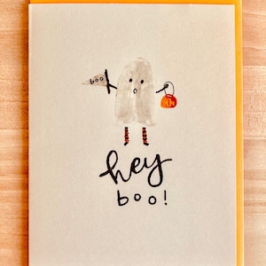 May include: A white ghost with black eyes and a surprised expression holds a small orange pumpkin bucket and a sign that says "boo". The ghost is wearing orange and black striped leggings. The text "hey boo!" is written below the ghost.
