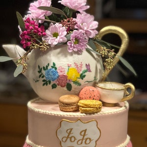 May include: A pink and white two-tiered cake decorated with a teapot, teacup, and macarons. The teapot is white with painted flowers and has a gold handle. The teacup is white with a gold rim. The macarons are pink and yellow. The cake is decorated with a gold sign that says "Jo Jo".