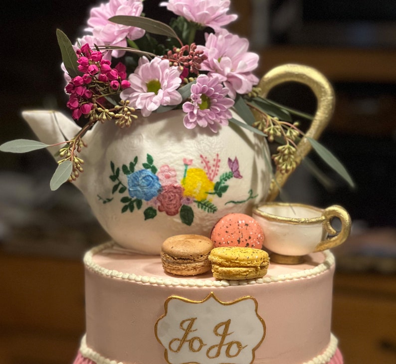May include: A pink and white two-tiered cake decorated with a teapot, teacup, and macarons. The teapot is white with painted flowers and has a gold handle. The teacup is white with a gold rim. The macarons are pink and yellow. The cake is decorated with a gold sign that says "Jo Jo".