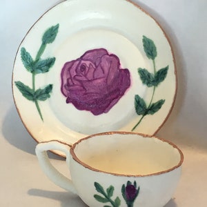 May include: A white teacup and saucer set with a hand-painted purple rose and green leaves. The set has a gold trim around the edges.