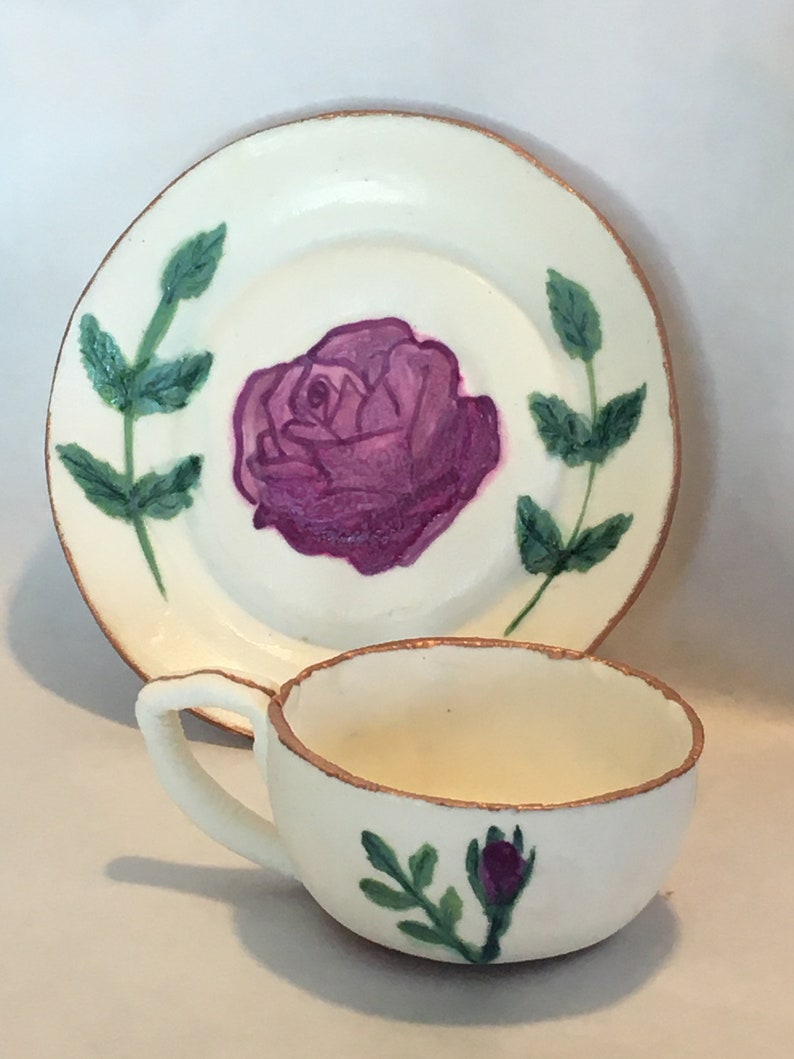 May include: A white teacup and saucer set with a hand-painted purple rose and green leaves. The set has a gold trim around the edges.