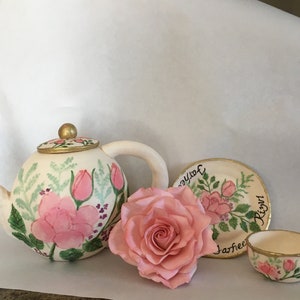 May include: A white teapot, teacup, and saucer with a gold rim, all decorated with pink roses and green leaves. The saucer has the text "Joy you" and "Jasheen Right" written on it. A single pink rose is also included.