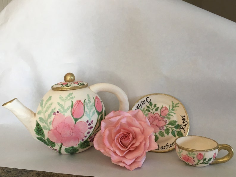 May include: A white teapot, teacup, and saucer with a gold rim, all decorated with pink roses and green leaves. The saucer has the text "Joy you" and "Jasheen Right" written on it. A single pink rose is also included.