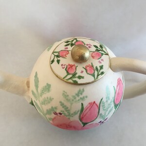 May include: A white teapot with a floral design. The teapot has a round body with a lid that has a gold knob. The floral design features pink flowers and green leaves.
