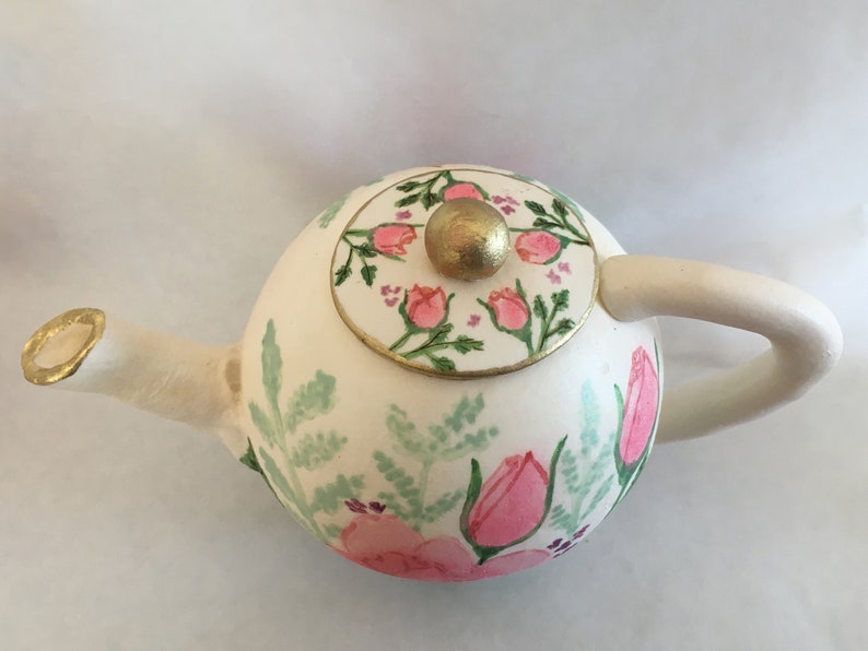 May include: A white teapot with a floral design. The teapot has a round body with a lid that has a gold knob. The floral design features pink flowers and green leaves.