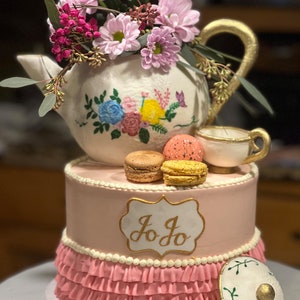 May include: A two-tiered pink cake decorated with a white teapot with floral designs, topped with pink and yellow macarons. The cake is decorated with a white ruffle and a gold label with the name "Jo Jo" written in cursive. The cake is on a white cake stand.