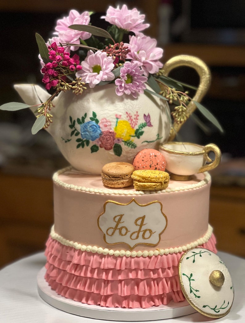May include: A two-tiered pink cake decorated with a white teapot with floral designs, topped with pink and yellow macarons. The cake is decorated with a white ruffle and a gold label with the name "Jo Jo" written in cursive. The cake is on a white cake stand.