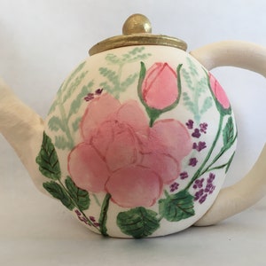 May include: A white ceramic teapot with a gold lid and a floral design. The teapot is painted with pink roses, green leaves, and purple flowers.