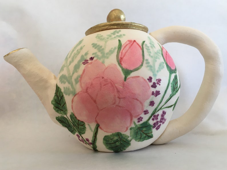 May include: A white ceramic teapot with a gold lid and a floral design. The teapot is painted with pink roses, green leaves, and purple flowers.