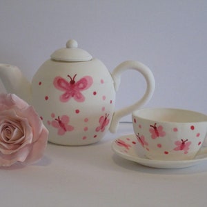 May include: A white teapot and teacup set with pink butterfly and polka dot designs. The teapot has a lid and a handle. The teacup has a saucer.