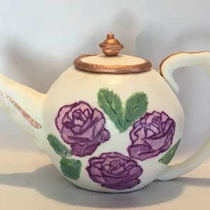 May include: A white teapot with a gold rim and a gold knob on the lid. The teapot is decorated with three purple roses and green leaves.