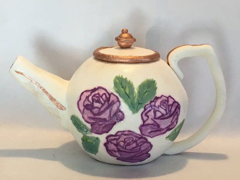 May include: A white teapot with a gold rim and a gold knob on the lid. The teapot is decorated with three purple roses and green leaves.