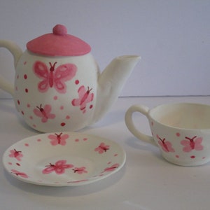 May include: A white tea set with pink butterfly and polka dot designs. The set includes a teapot, a cup, and a saucer.