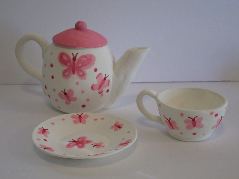 May include: A white tea set with pink butterfly and polka dot designs. The set includes a teapot, a cup, and a saucer.