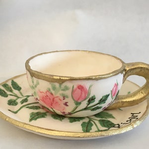 May include: A white teacup and saucer with a gold rim and floral design. The cup and saucer are painted with pink roses and green leaves. The saucer has the word "Biscotti" written on it.