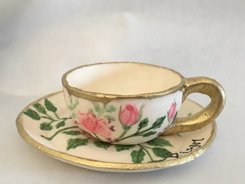 May include: A white teacup and saucer with a gold rim and floral design. The cup and saucer are painted with pink roses and green leaves. The saucer has the word "Biscotti" written on it.