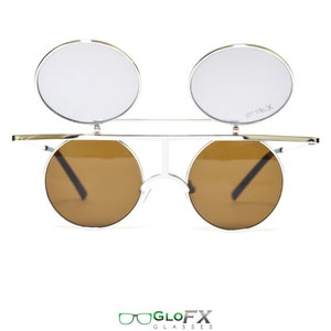 Op de afbeelding: Zilveren ronde zonnebril met bruine glazen en heldere omhoog te klappen glazen. De zonnebril heeft een metalen frame en de omhoog te klappen glazen hebben het woord "crotx" erop gedrukt.