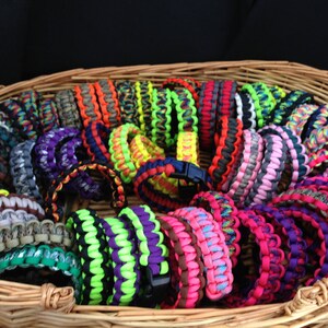 May include: A wicker basket filled with colorful paracord bracelets. The bracelets are made with different patterns and colors, including camouflage, neon green, pink, and blue.
