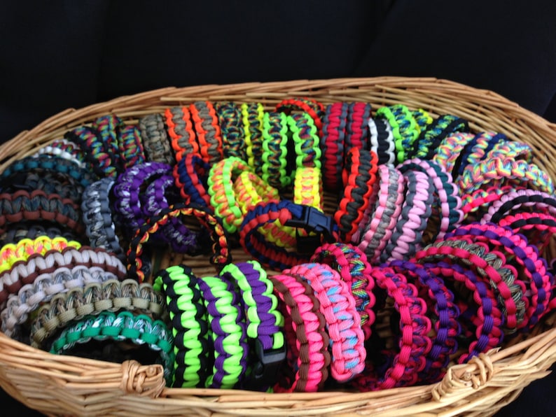 May include: A wicker basket filled with colorful paracord bracelets. The bracelets are made with different patterns and colors, including camouflage, neon green, pink, and blue.