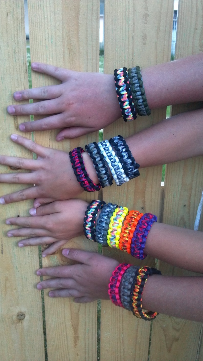 May include: A collection of colorful paracord bracelets with different patterns and colors, including black, white, green, yellow, red, and blue.