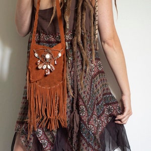 May include: A woman wearing a brown and white patterned dress with a brown suede fringe bag with a shell and bead embellishment. She is wearing shell anklets on both ankles.