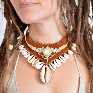 Könnte beinhalten: Eine braune Lederkette mit einem beigefarbenen Makrameemuster und weißen Muscheln. Die Kette hat einen türkisfarbenen Edelstein-Akzent.