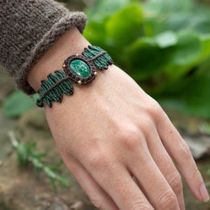 May include: A macramé bracelet with an oval green stone centre, framed by a brown border and small gold beads. The bracelet has a dark green leaf design on each side. The bracelet is on a wrist with a brown sleeve.