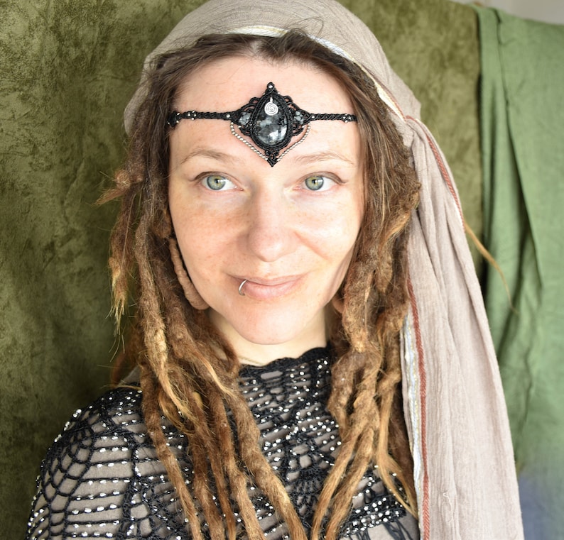 May include: A woman with long brown dreadlocks wears a black beaded headband with a large black gemstone pendant. The headband is adorned with silver accents. She is wearing a black crocheted top and a brown scarf.