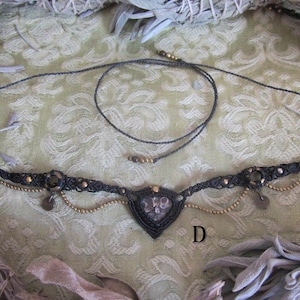 May include: A black macrame necklace with a heart-shaped pendant featuring a dark grey stone. The necklace has gold beads and charms.