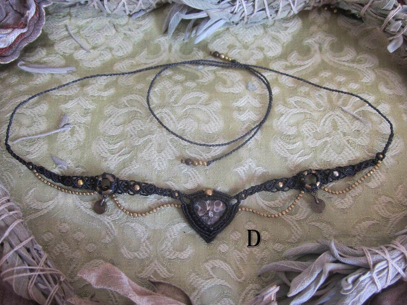 May include: A black macrame necklace with a heart-shaped pendant featuring a dark grey stone. The necklace has gold beads and charms.