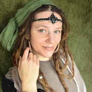 May include: A woman with long brown dreadlocks wears a green headscarf and a black beaded headband with a large black gemstone. She is smiling and looking at the camera.