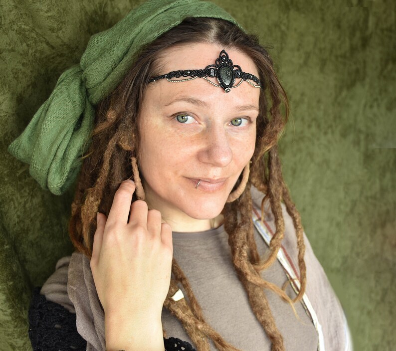May include: A woman with long brown dreadlocks wears a green headscarf and a black beaded headband with a large black gemstone. She is smiling and looking at the camera.