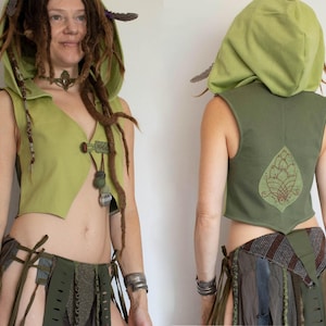 Könnte beinhalten: Eine ärmellose, hellgrüne Weste mit passender Kapuze. Die Weste hat einen Knopfverschluss und dekorative Akzente. Die Rückseite der Weste zeigt ein gesticktes Design. Das Outfit beinhaltet einen Rock mit mehreren Stoffstreifen.