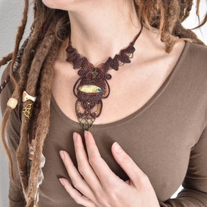 Op de afbeelding: Een bruine koordketting met een grote hanger van gele, groene en blauwe steen. De hanger is omgeven door ingewikkelde knoopwerk en een kleine, ronde metalen hanger.