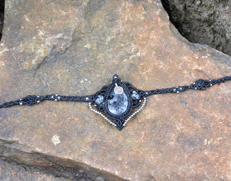 May include: A black macrame choker necklace with a silver pendant featuring a clear, gray, and black gemstone. The necklace is accented with silver beads.