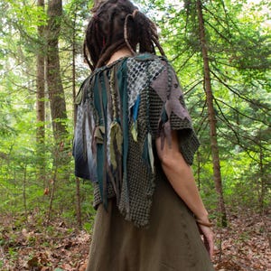 Könnte beinhalten: Ein strukturierter, olivgrüner Strickponcho mit geschichteten Stoffstreifen, Netzpaneelen und hängenden Fransen. Das Kleidungsstück wird über einem einfarbigen, olivgrünen Kleid getragen und zeigt eine Mischung aus Strickmustern und Stoffen.