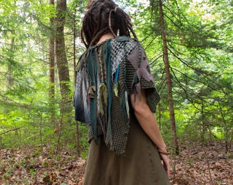 Peut inclure: Un poncho en tricot vert olive texturé avec des bandes de tissu superposées, des panneaux en maille et des détails à franges pendantes. Le vêtement est porté sur une robe vert olive unie, présentant un mélange de mailles et de textures.