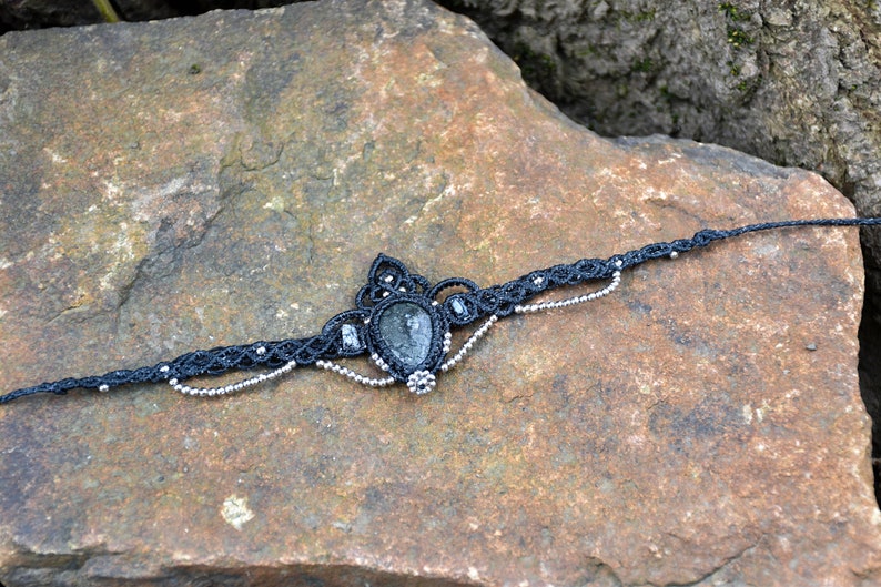 May include: A black macrame choker necklace with a dark green gemstone pendant. The necklace is adorned with silver beads.