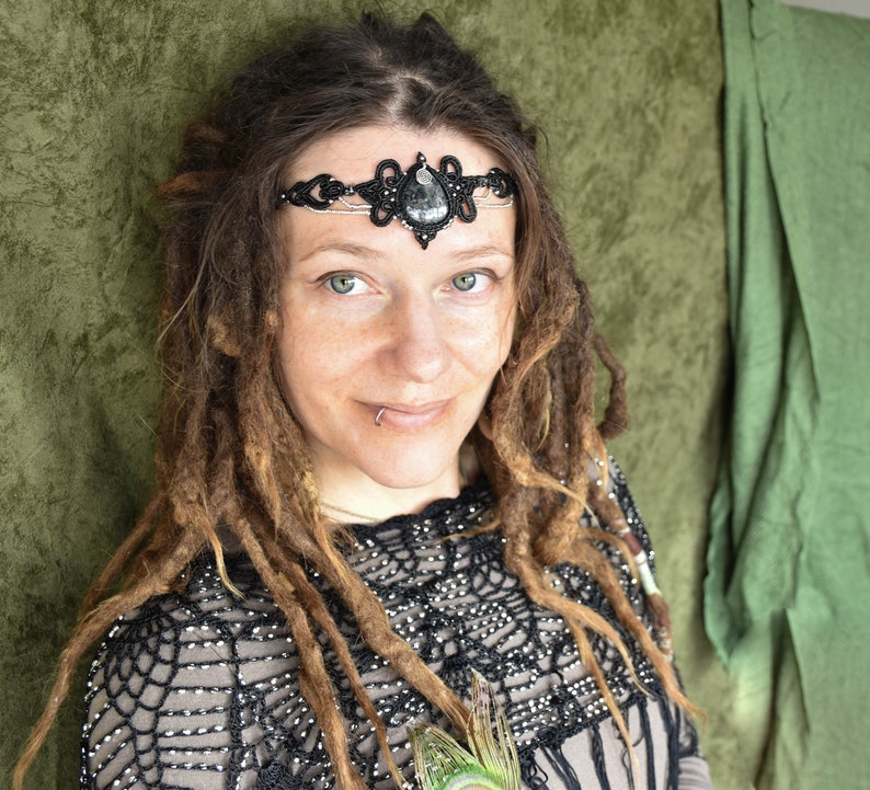 May include: A woman with long brown dreadlocks wears a black beaded headband with a large black gemstone. The headband is adorned with intricate silver wire details. She is wearing a black and silver crochet top.