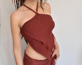 May include: A rust-colored halter top featuring a textured, layered fabric design with a pointed hemline. The garment has thin straps that tie around the neck and is paired with a matching rust-colored wrap skirt that has a frayed, fringed hemline.