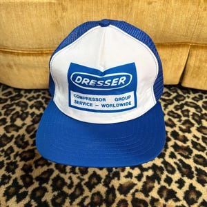 May include: A blue and white trucker hat featuring a mesh back and a solid front panel. The front displays a blue rectangular logo with the text DRESSER COMPRESSOR GROUP SERVICE - WORLDWIDE printed in white and blue lettering on a white background.