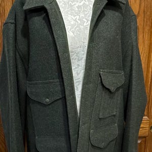 May include: Dark green wool coat with four front pockets and a notched collar. The coat is displayed on a mannequin against a wooden door background. The coat has a button closure.