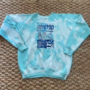 May include: A light blue and white tie-dye sweatshirt with the text "The Pajama Game" and event details in blue. The raglan sleeves and crew neck are visible. The sweatshirt is laid flat on a textured surface.