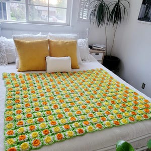 May include: A crocheted throw blanket with a daisy pattern in shades of green, yellow, and orange. The blanket is on a white bed with yellow and white pillows.